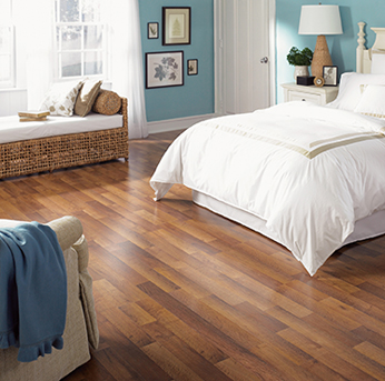 A bright bedroom with warm, medium-toned natural hardwood flooring, a white-clad bed, and a decorative woven bench